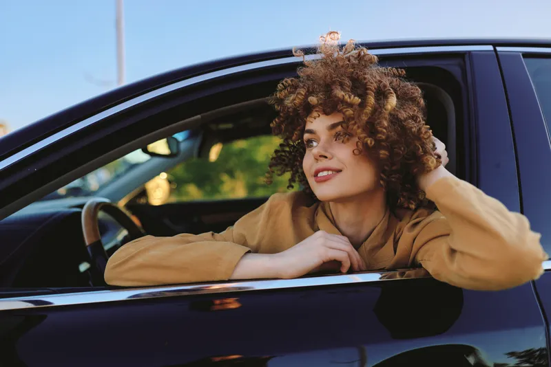 Cheerful woman leaning from a car window, relaxed pose, sunlit morning, casual sweater, soft breeze, retro film color palette, natural tones, candid lifestyle moment