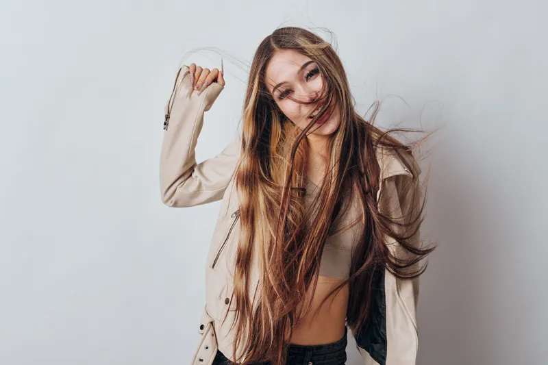 Young woman with long hair smiling and posing in casual jacket and crop top against white studio background for fashion portrait.