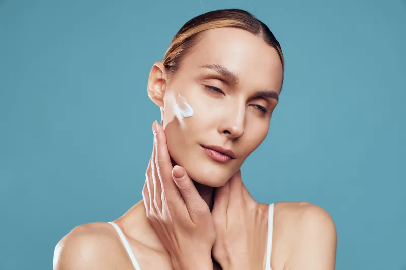 Young woman with fair skin applies moisturizing cream on her cheek while standing against a blue background with closed eyes and a calm expression.