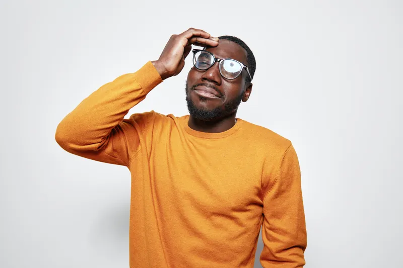 African American man with dark skin wearing glasses and orange sweater touching head with thoughtful expression on white background.