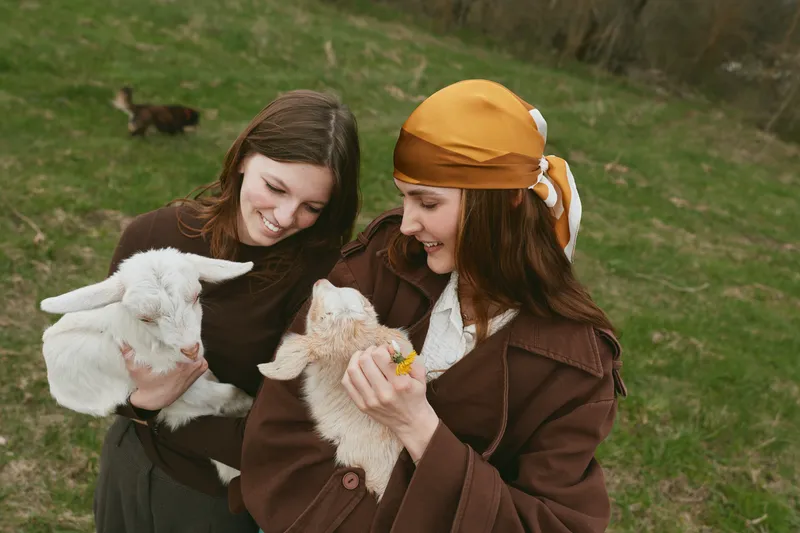 Two women outdoors cradle fluffy goat kids, sharing smiles as they interact with playful animals. Natural daylight on a grassy field creates a warm, friendly lifestyle moment.