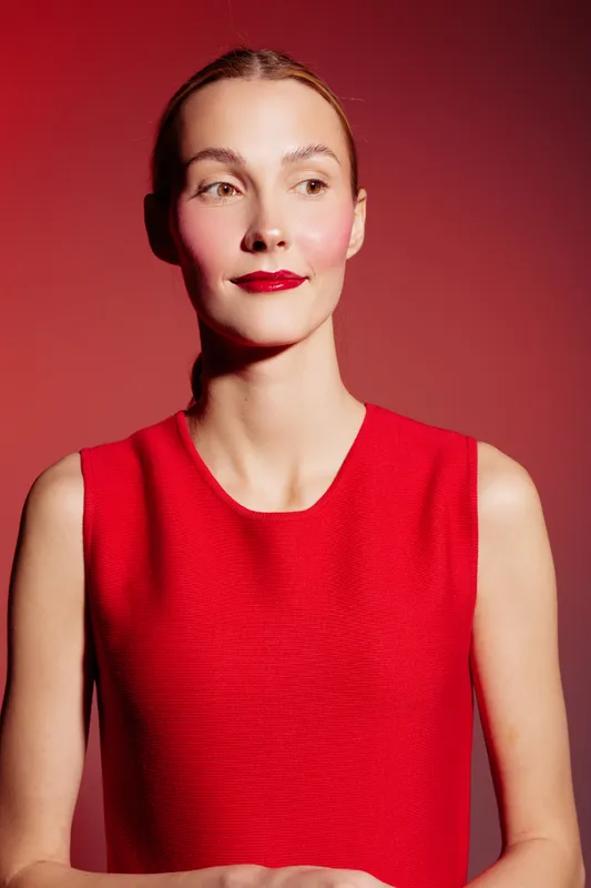 Young woman in red sleeveless dress poses against red background with confident expression. Studio portrait with bright lighting and minimalistic style. Concept of fashion and elegance.