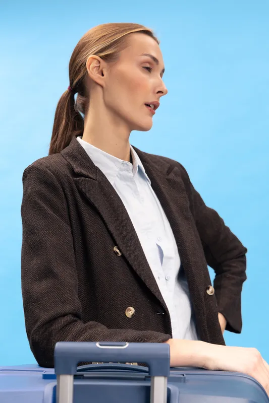 Young woman in brown blazer stands with hand on hip near suitcase on blue background. Concept for travel, business trip, and stylish female traveler with confident posture and calm expression.