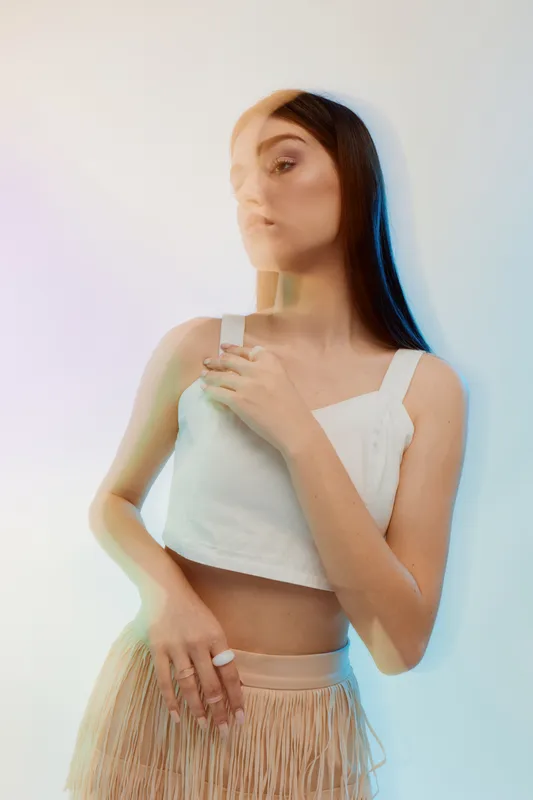Young woman with long dark hair poses in a white top and beige skirt with fringe. Soft lighting and blurred effect create a dreamy atmosphere. Concept of beauty fashion style.