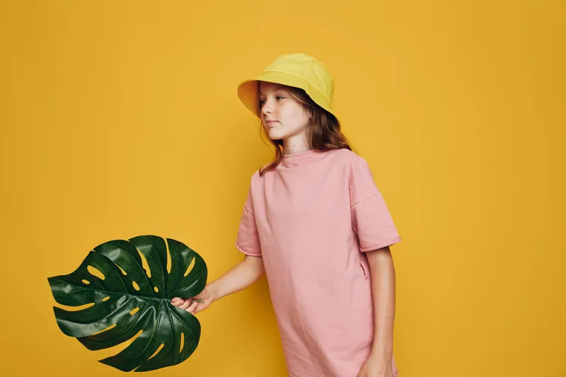 Girl in yellow hat and pink shirt holds large green leaf against yellow background. Studio portrait with natural light. Concept of nature and childhood.
