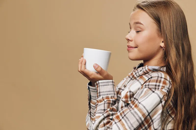Girl holding cup with both hands close to face smelling aroma enjoying hot drink with eyes closed in cozy plaid shirt standing on beige background.
