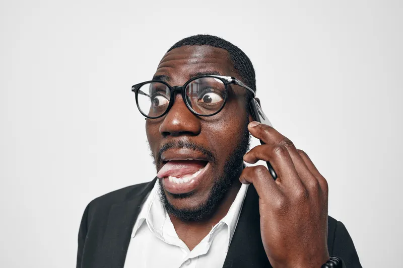 African American man with dark skin and glasses talking on smartphone with surprised expression in studio. Casual business attire and white background for communication concept.