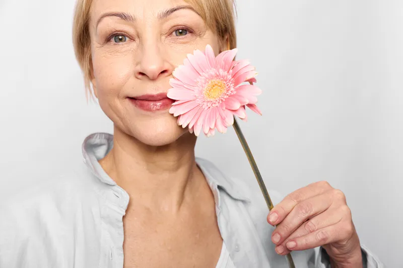 Smiling woman holds a pink flower near her lips in a bright closeup, beauty portrait with skincare and wellness mood, soft studio light, gentle self care