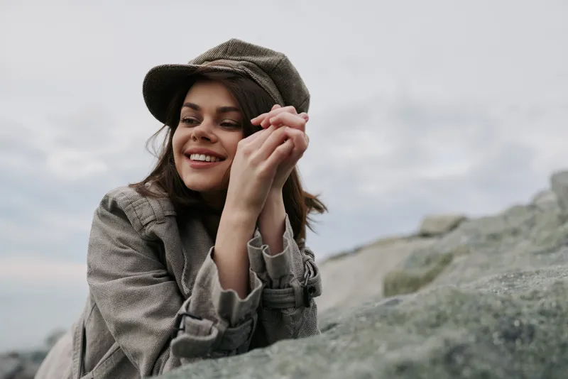 Smiling young woman in a brown plaid coat and hat leans on a rock outdoors under cloudy sky. Natural light highlights her joyful expression and casual autumn fashion style.