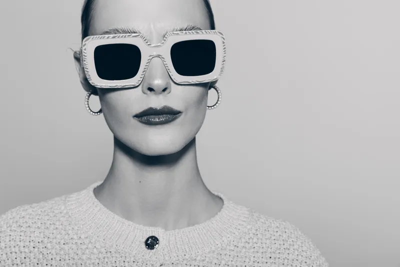A pale Caucasian woman with smooth skin wears oversized white sunglasses and hoop earrings. Monochrome portrait with a minimalist style and neutral expression.