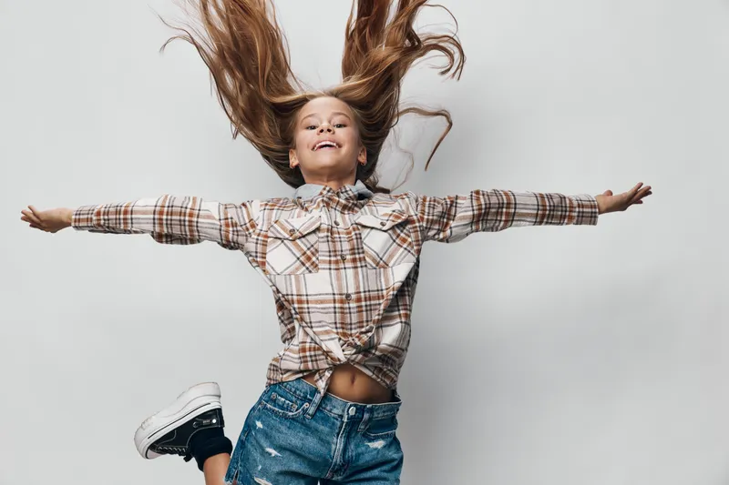 Girl jumping with arms outstretched wearing checkered shirt and denim shorts on white background smiling with hair flying for energetic joyful motion and freedom.