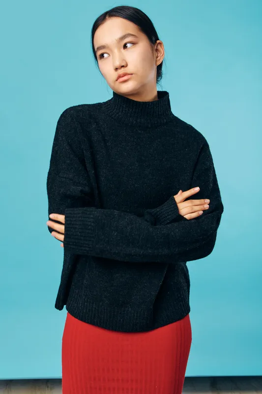 Young woman with crossed arms wearing black sweater and red skirt standing against blue background looking sideways with serious expression. Casual fashion and portrait concept.