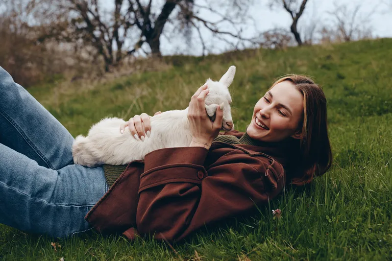 A joyful goat kid rests on a grassy field with a smiling woman outdoors, relaxation, bonding, casual sweater. A lifestyle scene highlighting nature, countryside moments and calm companionship.