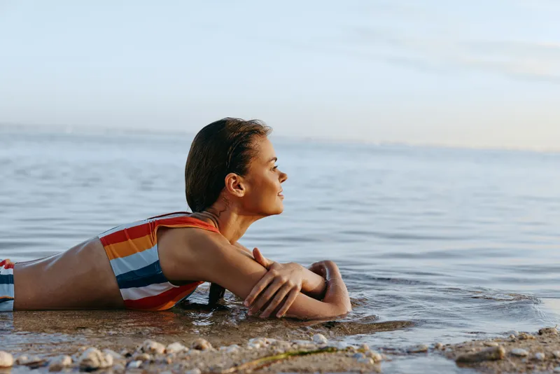 Young woman ocean beach shallow water lying swimsuit, striped swimwear, resting on wet rocks at shoreline, calm summer morning, side view, relaxed expression, copy space, peaceful waves