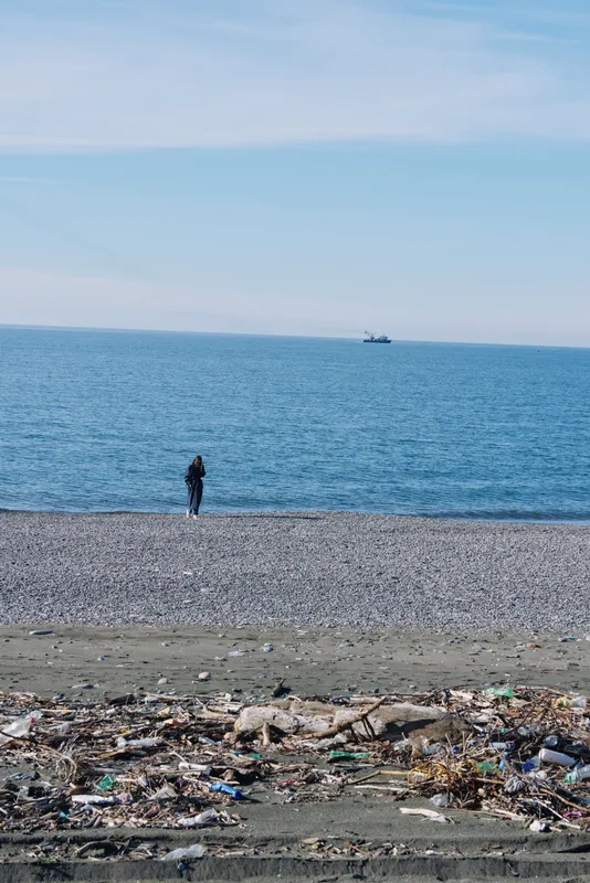 Beach scene with a lone person walking along a pebbled shoreline, highlighting pollution and litter at the water edge, with the sea on the horizon.
