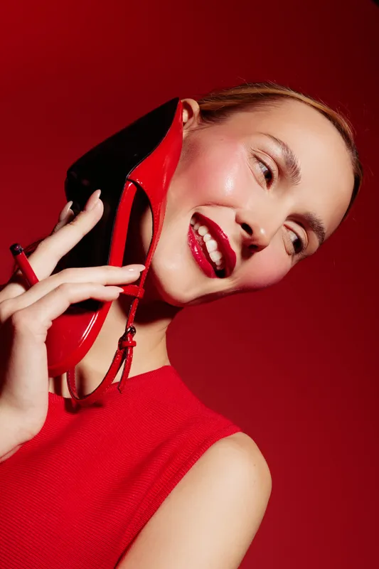 Smiling woman in red dress holds red high heel shoe near ear like a phone, posing on red background. Concept for fashion, style, beauty, and creative advertising with vibrant colors.