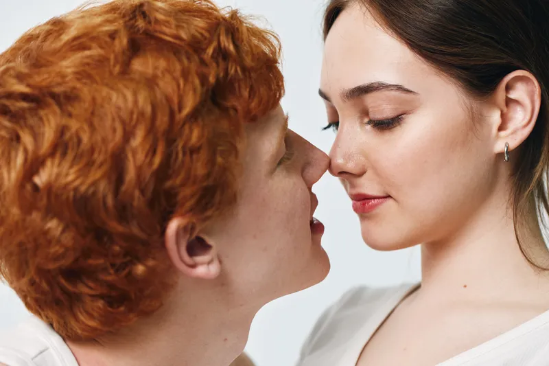 Young couple with red hair and brunette face about to kiss in closeup, romantic moment with closed eyes and soft expression on light background for love and intimacy.