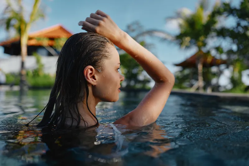 Cheerful woman in a pool outdoors, sunlit water and relaxed posture, wet hair against bright blue reflections, tropical ambiance, natural colors, carefree lifestyle