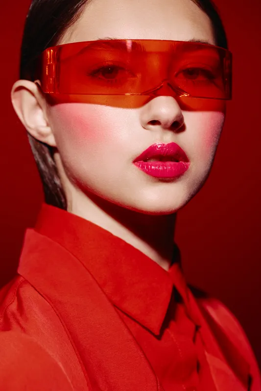 Fashion woman with red lipstick and blush wearing translucent red sunglasses and bright clothing. Creative beauty portrait on red background. Concept for bold makeup and style.