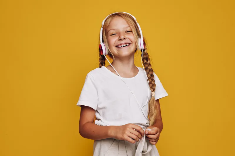 Happy girl with blond hair in braids wearing headphones and white t-shirt smiles while standing against a yellow background. Studio portrait of cheerful child.
