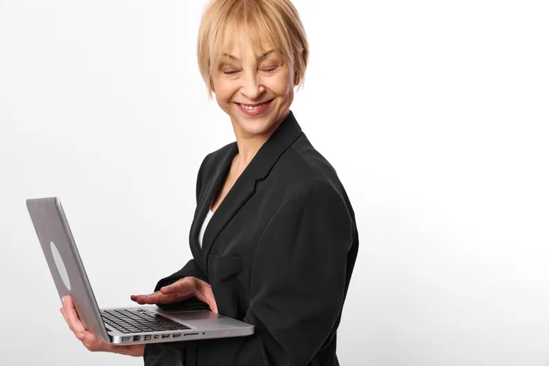Smiling businesswoman, senior woman with laptop, standing in studio, black suit jacket, focused hands typing, clean neutral background, copy space