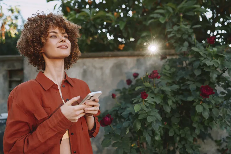 Curly haired woman in a rust shirt stands in a garden, smiling while checking a smartphone. Warm daylight highlights foliage, creating a relaxed lifestyle mood and warmth