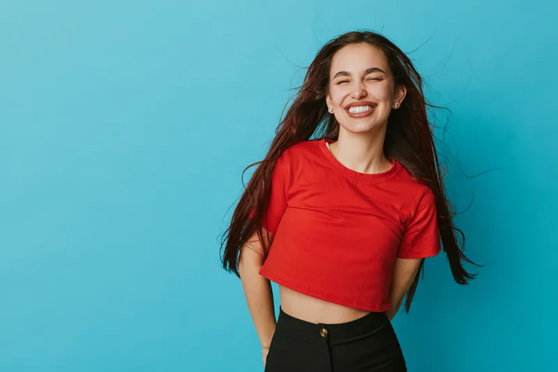 Young woman smiling with eyes closed wearing red crop top and black pants standing against blue background. Happy female with long hair enjoying moment in casual outfit with joyful expression.