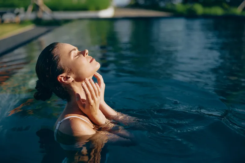 Woman in a sunlit pool, eyes closed, enjoying warm water and a gentle breeze, relaxed posture in an outdoors setting, sunlight on skin, vibrant colors and serene mood, refreshing moment of leisure