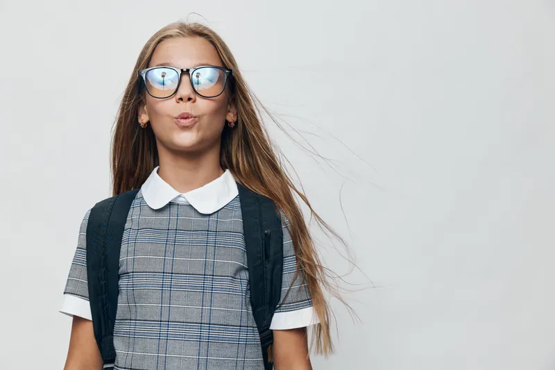 Girl with glasses and backpack standing in studio with blowing hair, wearing checkered dress with white collar, isolated on white background for school or education concept.