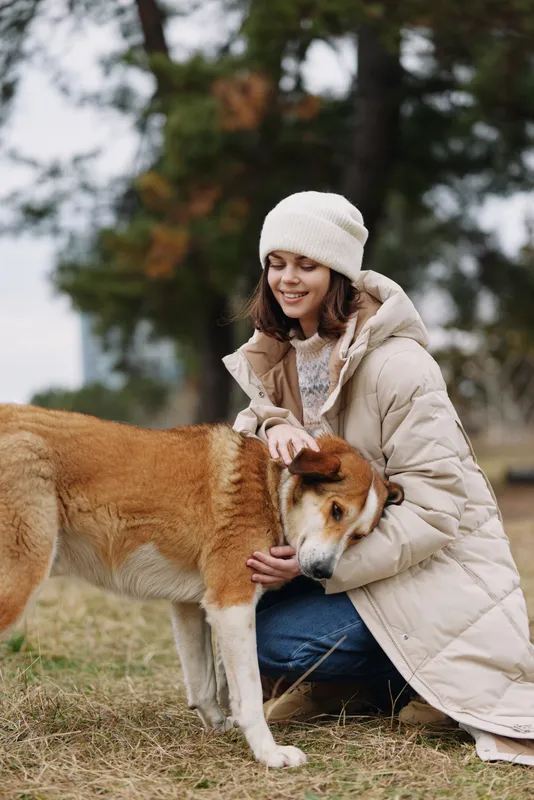 Woman kneels in a warm coat and beanie, gently petting a dog in an outdoor park setting with autumn light, capturing calm companionship and care in nature