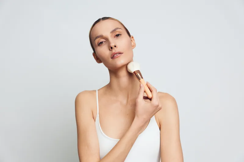 A young woman in a white tank top applies makeup with a brush on her neck against a plain light background. The concept focuses on skincare and beauty routine.
