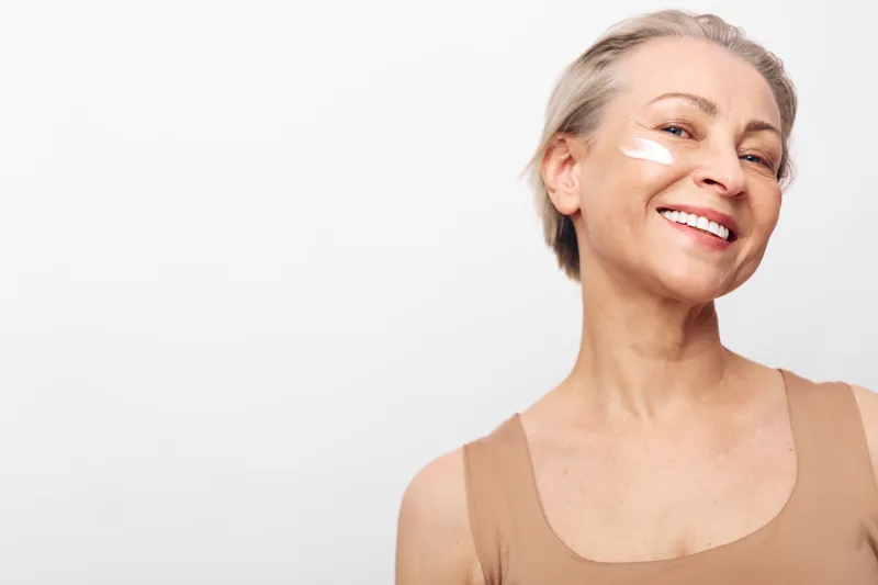 Smiling mature woman with short gray hair applies white facial cream on cheek. Close-up portrait with clear skin in beige top on white background. Concept of skincare, beauty, and self-care.