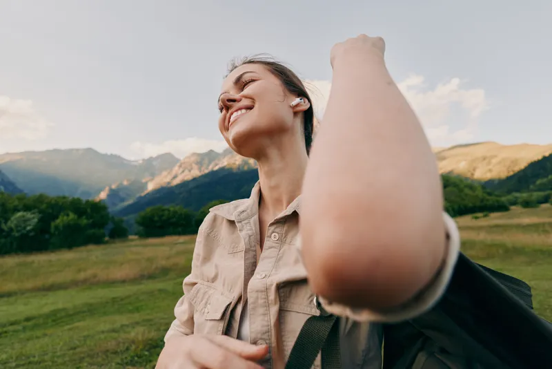 Cheerful woman outdoors, smiling, backpack ready, nature surrounds a scenic field with distant mountains, daylight glow, casual style, relaxed pose, joyful moment in open landscape