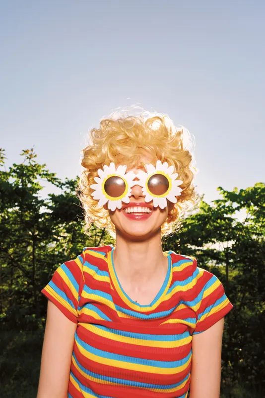 Cheerful woman outdoors, wearing flower sunglasses and striped tee, sunlit moment, carefree mood, vibrant colors, lush greenery background