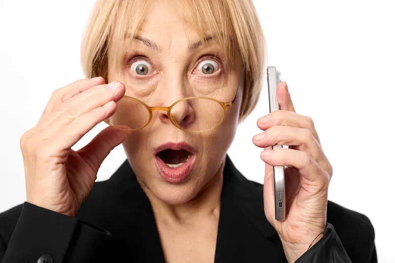 Surprised blonde woman holding novelty glasses near her eyes and a smartphone in one hand, studio portrait on white background, wide eyed expression, humor and communication theme