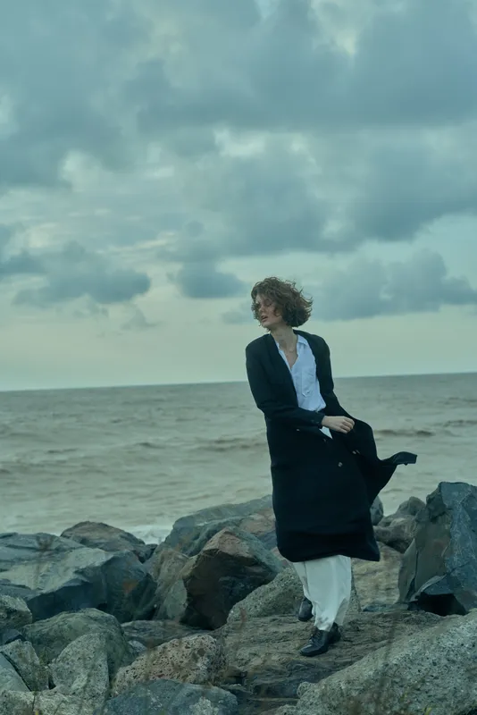 Sea breeze on a rocky coast, a woman in a long coat stands on stone by the shore, contemplative mood, muted tones, windy weather, minimalist outdoor fashion scene by the ocean