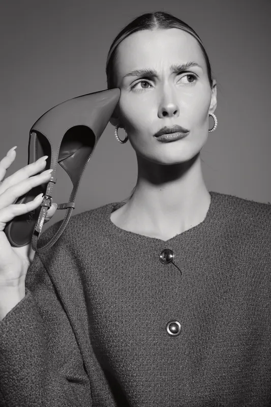 Serious Caucasian woman with pale skin holds high heel shoe near face, wearing earrings and textured coat. Studio portrait in black and white.