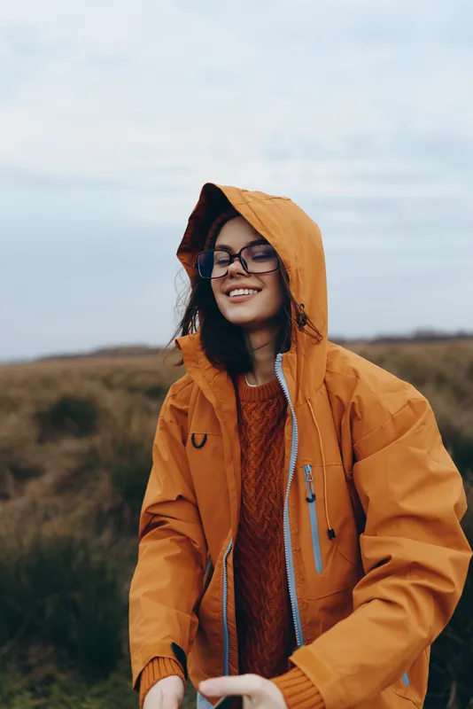 Woman lifestyle outdoors portrait, smiling woman in hooded orange coat, wearing glasses and scarf, standing on windy autumn hillside, soft daylight, relaxed pose, natural colors, calm countryside