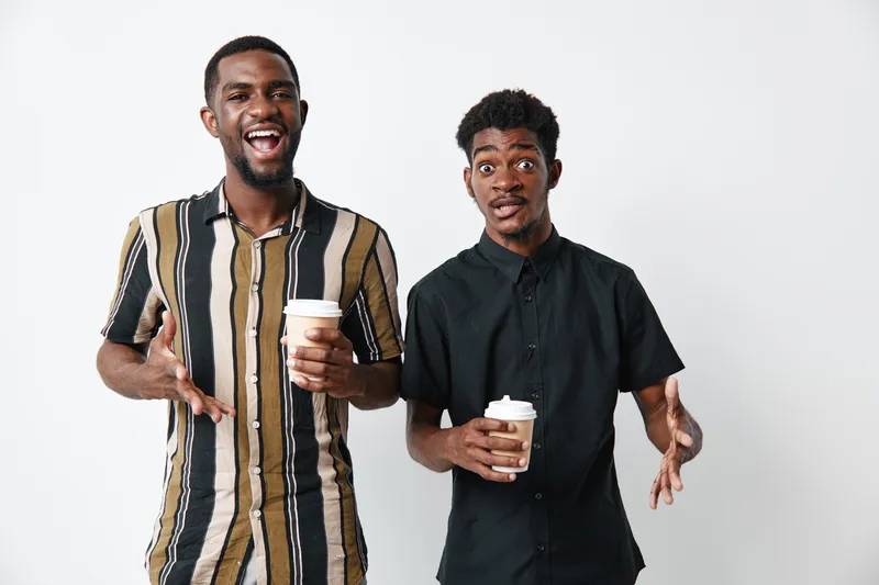 Two young black men with dark skin holding coffee cups and gesturing while standing against white background. Casual clothing and friendly expressions in studio setting.
