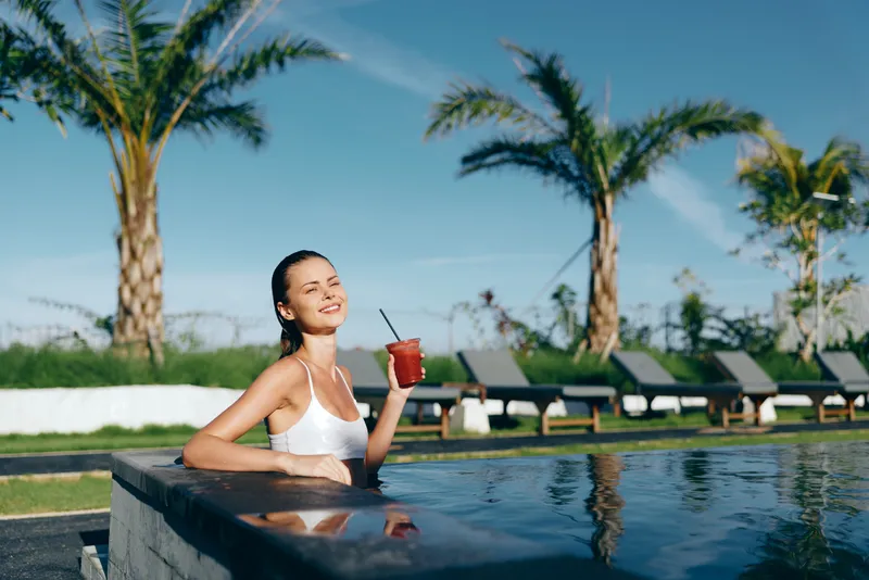Smiling woman in white bikini relaxing in outdoor pool holding a red cocktail with straw. Palm trees and lounge chairs in background under clear blue sky. Lifestyle and summer concept.