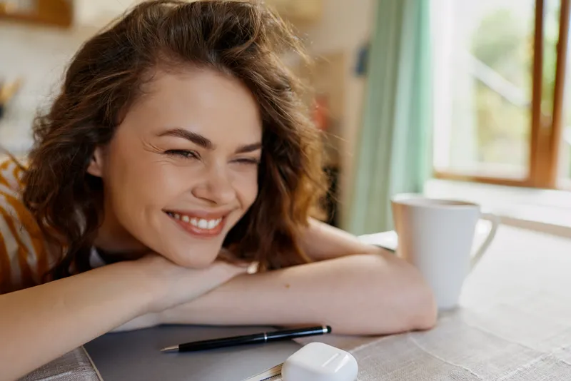 Smiling woman rests her chin on folded arms at a home desk, warm natural light from a window, mug nearby, pen and notebook suggesting casual work and lifestyle.
