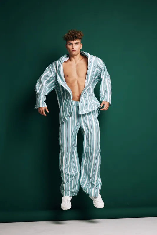Young man jumping in striped loose suit with open shirt and white sneakers on green background. Fashion model with curly hair posing in studio with casual outfit and confident expression.