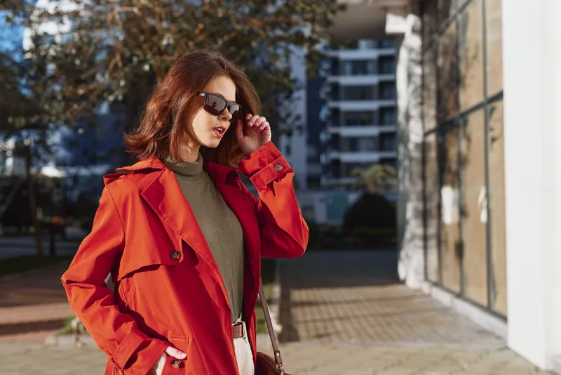 Stylish woman wearing red coat and sunglasses walking outdoors in urban setting, sunny day, confident expression, modern casual outfit, autumn season