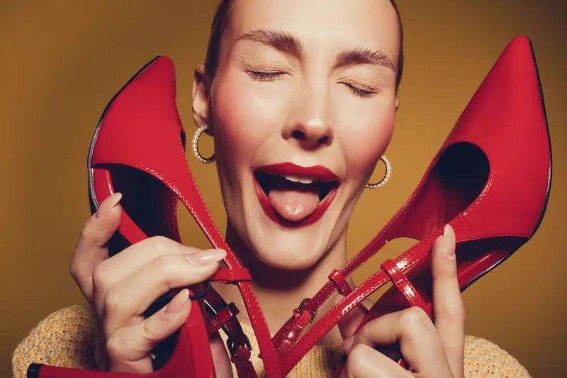Happy woman with closed eyes holds red high heels near face, smiling with tongue out. Bright studio lighting highlights joyful mood and vibrant fashion concept with stylish footwear.