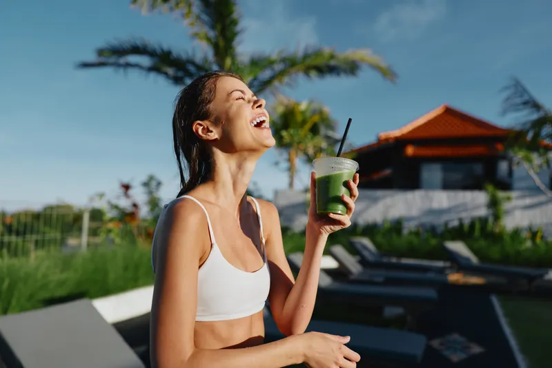 Cheerful woman outdoors by poolside, sunlit smoothie drink moment, fitness vibe, relaxed posture, breeze, vibrant colors, wellness focus, summer mood, casual lifestyle, vacation backdrop, healthy