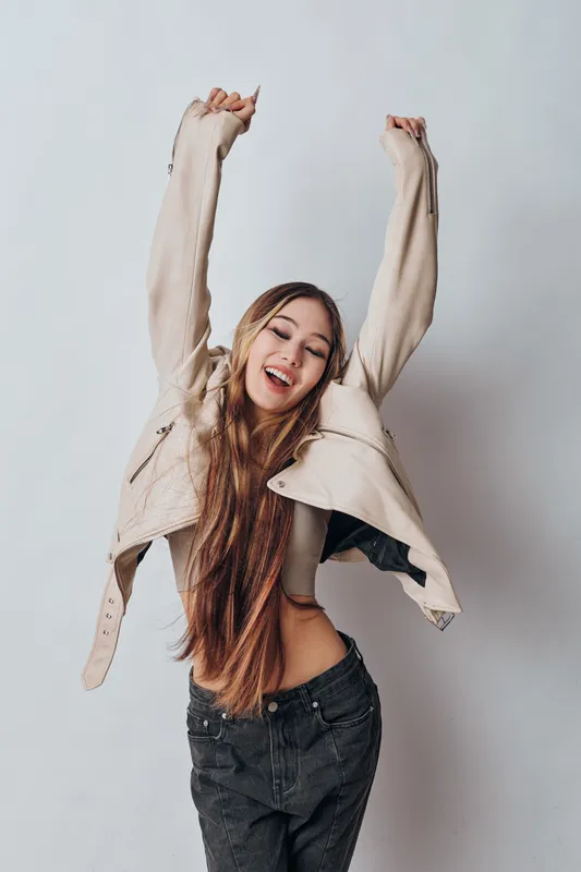 Young woman with long hair smiling and stretching arms up wearing beige jacket and dark pants, posing happily in studio against plain light background.