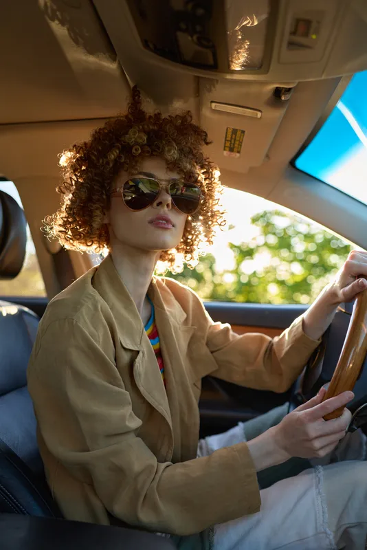 Woman driver sits behind the wheel with curly hair and sunglasses in a retro fashion style, inside a car bathed in warm light, showcasing a casual lifestyle moment