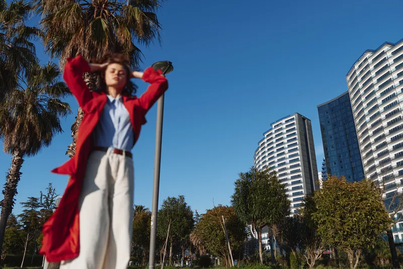 Woman in a red coat and white pants stands outdoors in sunny urban park with arms raised, city skyline behind her, vibrant mood and fashion forward attitude