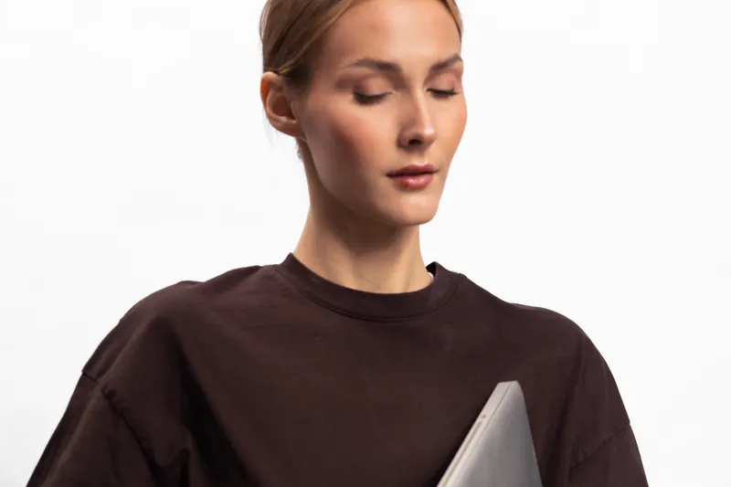 Young woman with closed eyes holds laptop close to chest wearing dark brown sweater. Studio portrait on white background. Concept of focus, calm, technology and casual style.