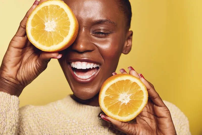 Happy African woman with dark skin holds two orange halves near her face smiling with closed eyes on yellow background. Concept of healthy lifestyle, vitamin C, and natural beauty.